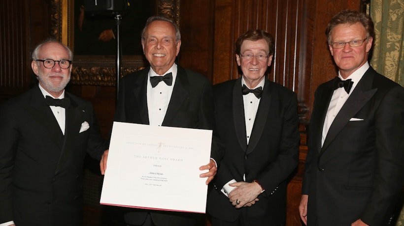 Four people wearing black tuxedos facing forward. One of them is holding an award certificate.