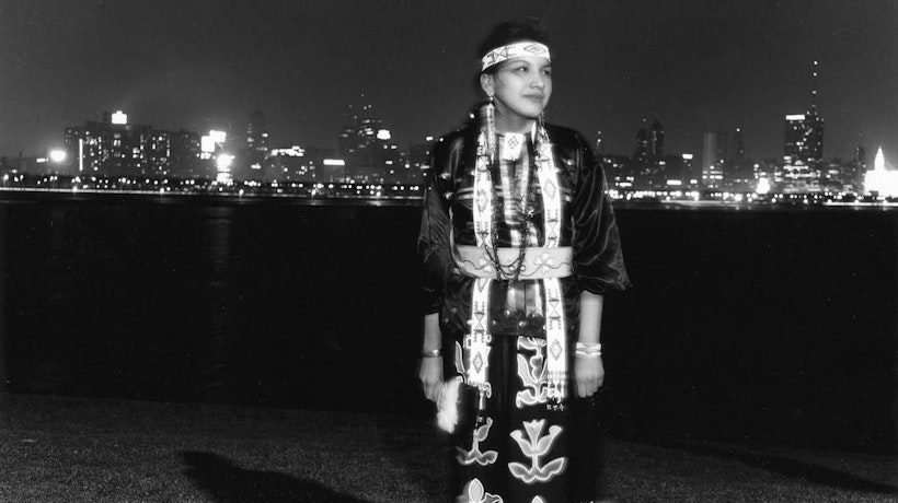 A black and white photograph of a person wearing native clothing standing with an urban landscape behind them.